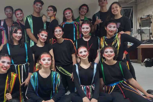 Grupo de Teatro do colégio Objetivo reunidos em três fileiras. Todos estão vestindo figurinos com roupas pretas e fitas coloridas presas aos ombros. Estão maquiados com pintura colorida em volta dos olhos e o rosto pintado de branco.