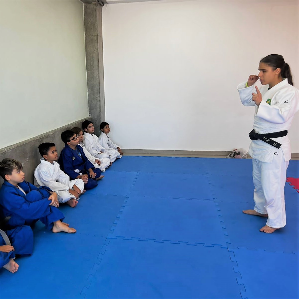 Atleta de Judô orientando os alunos do colégio Objetivo sobre a importância de estimular os sentidos sensoriais do corpo, na imagem os alunos estão sentados a esquerda em fila e a atleta a direita e em pé está com sua mão direita próxima a orelha indicando para usar a audição no treino.