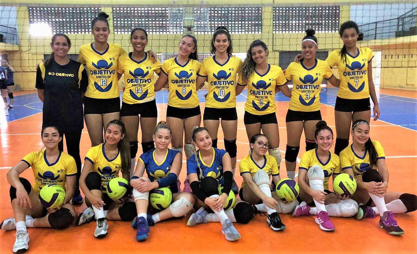 Time feminino de Vôlei do Colégio Objetivo reunido em duas fileiras. Fileiras de trás as atletas estão de pé e na fileira da frente estão sentadas no chão. A rede da quadra está de fundo na imagem.