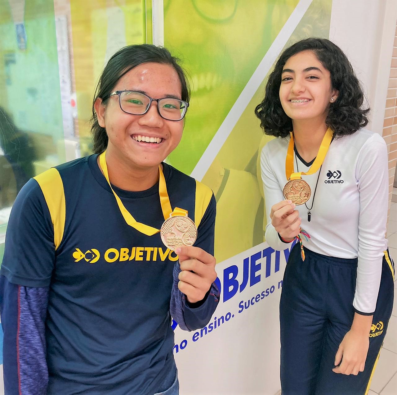 Alunos medalhistas ilustram o GAP Olímipico do colégio Objetivo. Na foto temos um aluno a esquerda segurando sua medalha e a direita uma aluna segura a sua. Ambos vencedores de olimpíadas de conhecimento.