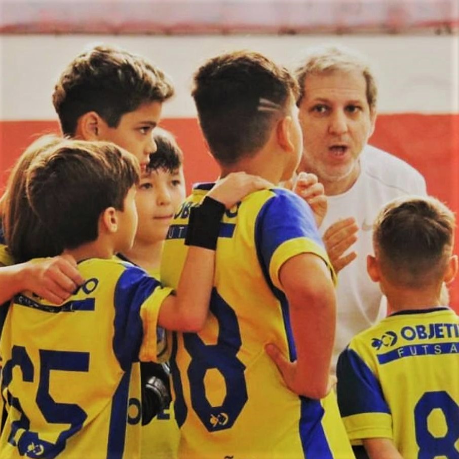 Time de Futsal Masculino do colégio Objetivo em um momento onde o técnico dos atletas passa as orientações estratégicas de jogo. Estão formando uma espécie de circulo em volta dele e apresentam estar focados em ouvir o professor na imagem.